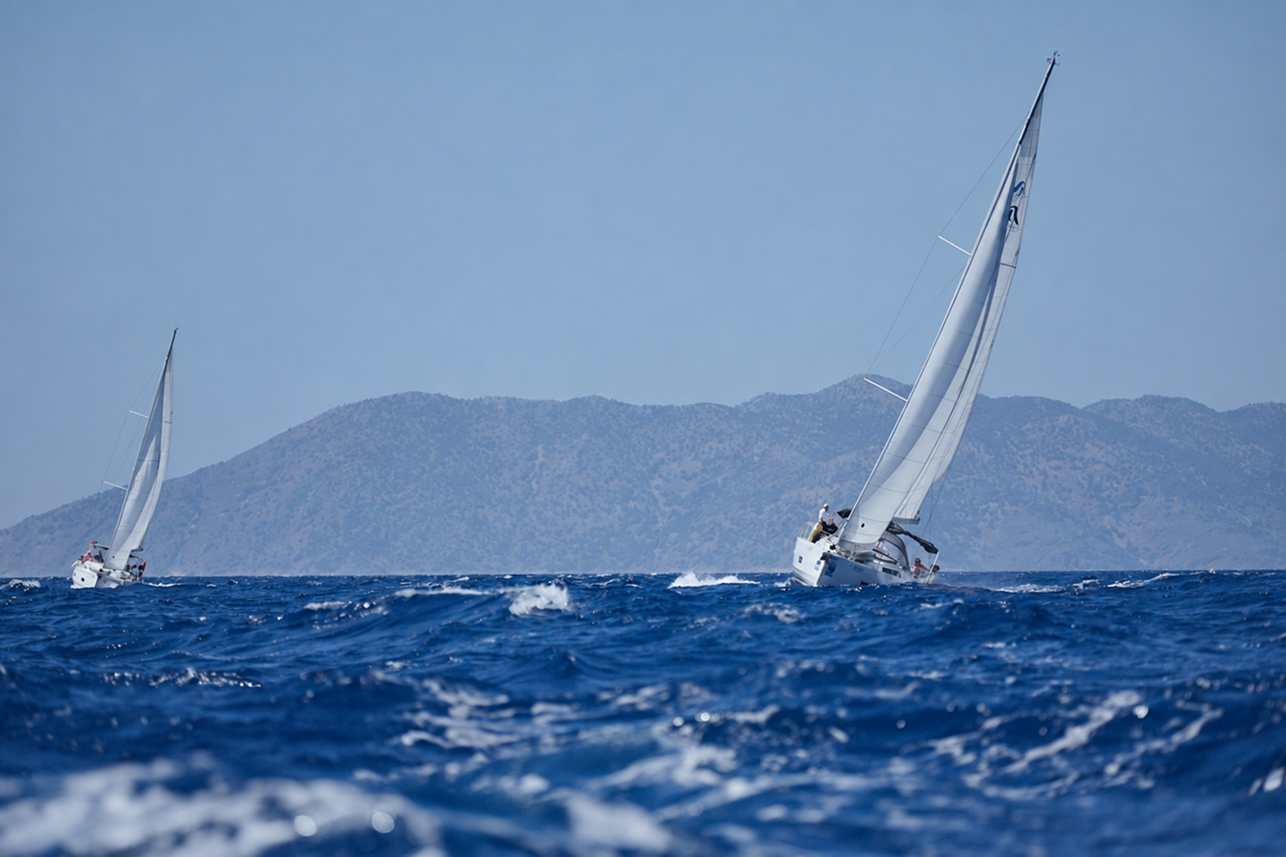 Two sailing yachts crossing blue water in front of a mountainous coastline